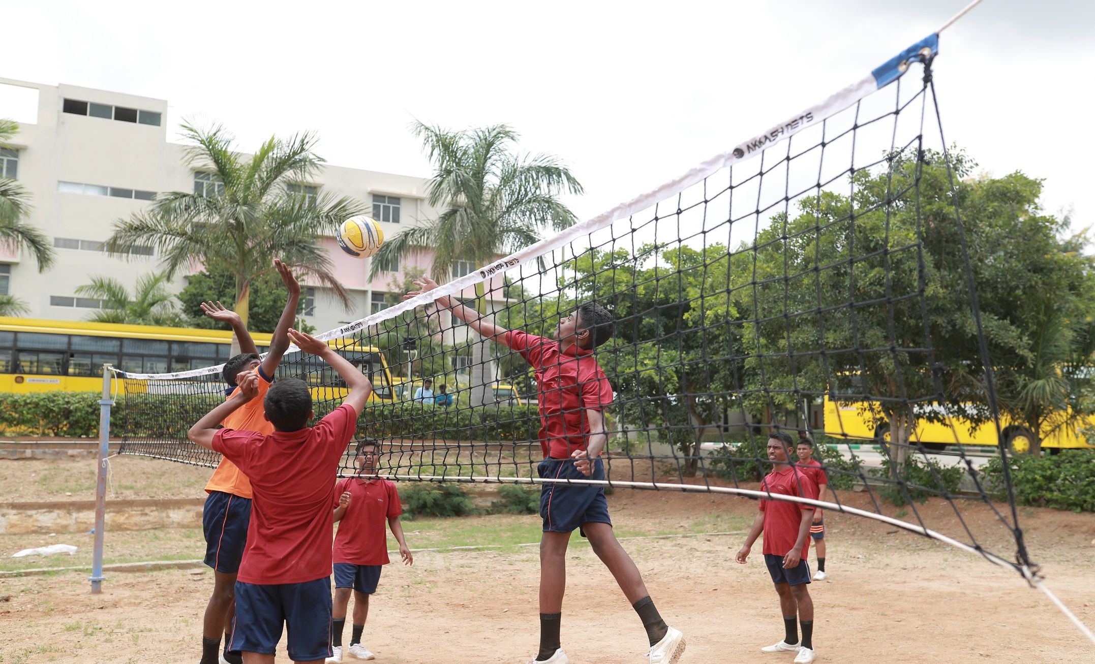 A player in a red shirt leaping to spike a volleyball over the net while teammates look on, palm trees and a yellow bus behind